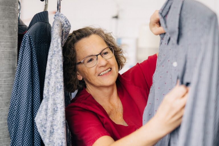 Eine Frau mit Brille und rotem Oberteil sortiert Hemden an einer Kleiderstange und lächelt dabei.