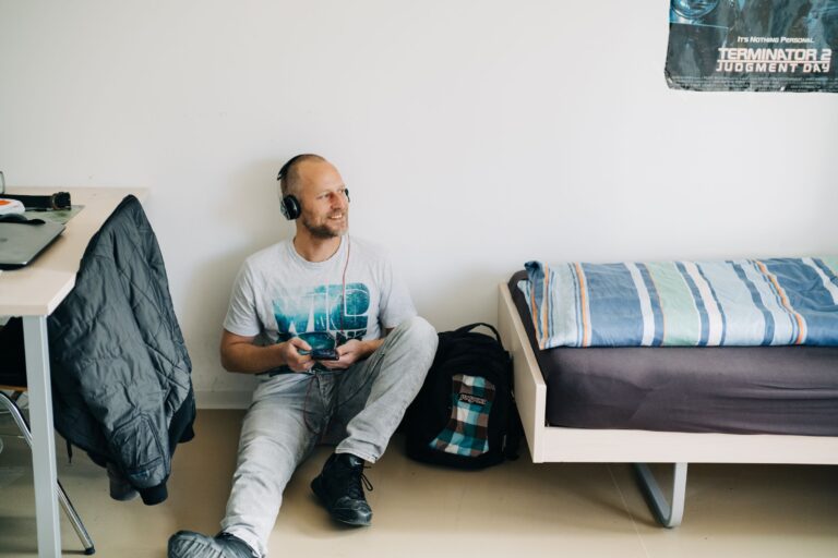 Mann mit Glatze und Kopfhörern sitzt entspannt am Boden eines schlichten Schlafzimmers neben einem Einzelbett und einem Rucksack, lächelt und hält ein Smartphone in der Hand. An der Wand hängt ein Filmplakat.