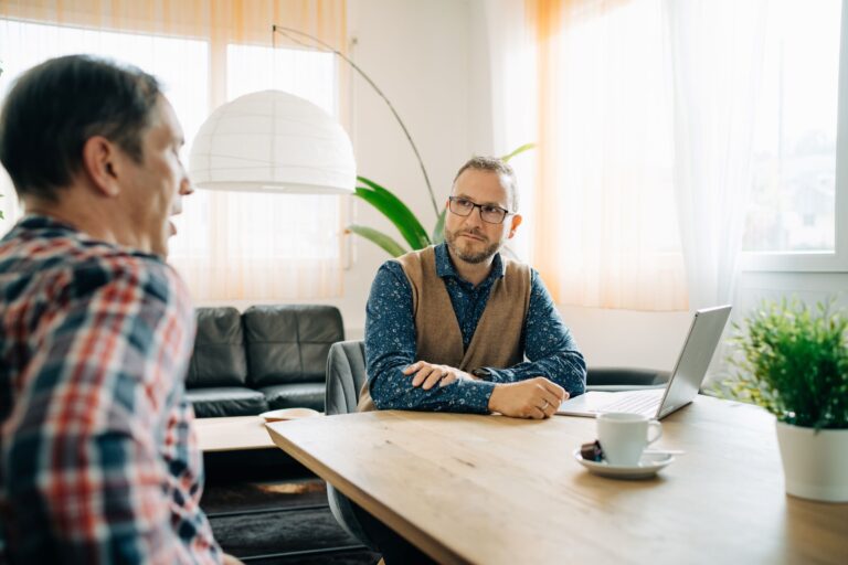 Mann mit Brille und Weste sitzt an einem Holztisch in einem sonnendurchfluteten Raum, hört aufmerksam zu und schaut seinen Gesprächspartner an. Im Hintergrund sind ein Sofa, eine Stehlampe und grosse Fenster mit hellen Vorhängen sichtbar.