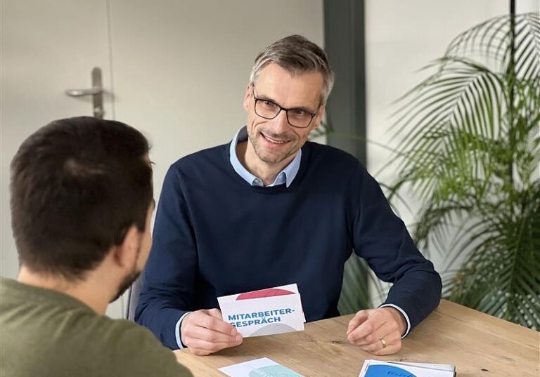 Mann mit Brille und dunkelblauem Pullover sitzt an einem Tisch und hält eine Karte mit der Aufschrift „Mitarbeitergespräch“ in der Hand. Er lächelt und spricht mit einer anderen Person, im Hintergrund stehen Pflanzen.
