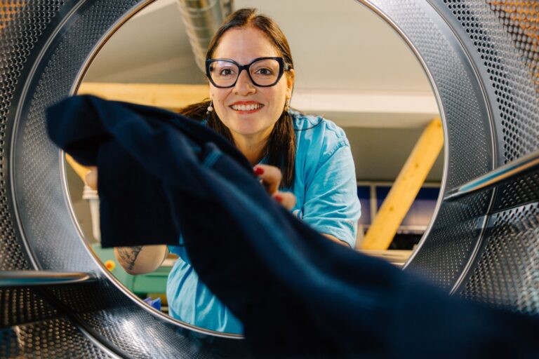 Frau mit Brille und türkisfarbenem Shirt lächelt und befüllt eine Waschmaschine, fotografiert aus der Perspektive des Trommelinneren.