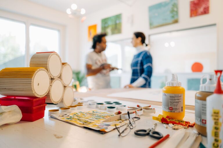 Heller Atelierraum mit Bastelmaterialien wie Farben, Klebstoff, Schere und Kartonrollen auf einem Tisch im Vordergrund. Im Hintergrund unterhalten sich zwei Personen, an der Wand hängen mehrere farbige Bilder.