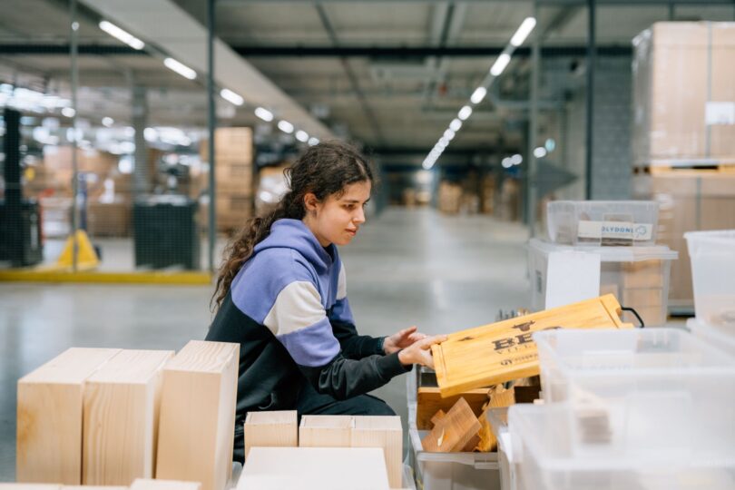 Junge Frau mit langen dunklen Haaren und Kapuzenpullover arbeitet in einer grossen Lagerhalle. Sie sortiert Holzteile und hält eine gelbe Holzkiste mit der Aufschrift „BBQ“ in der Hand. Um sie herum stehen weitere Holzteile und Plastikboxen.