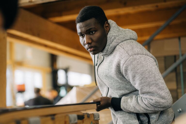 Junge Person mit grauem Hoodie arbeitet konzentriert an einer Werkbank in einer Werkstatt mit Holzbalken und Treppe im Hintergrund.