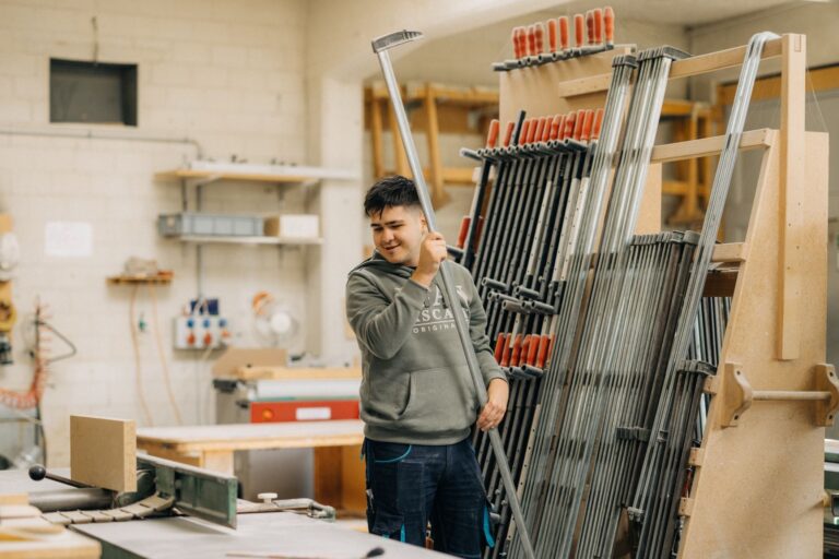 Junge Person in einer Werkstatt, umgeben von vielen Metallstangen und Werkzeugen, hält eine lange Metallstange in der Hand und schaut lächelnd zur Seite.