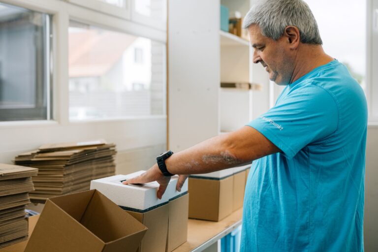 Mann mit grauen Haaren und türkisfarbenem Shirt verpackt Schaumstoffteile in Kartons an einem Arbeitstisch neben einem Fenster. Im Hintergrund sind gestapelte Kartons sichtbar.