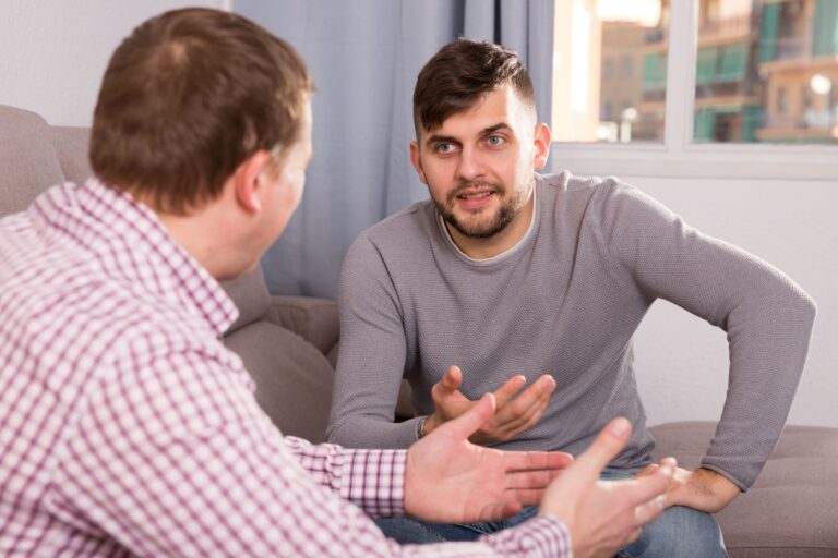 Zwei Männer sitzen auf einem Sofa und führen ein intensives Gespräch. Der Mann im Vordergrund trägt ein kariertes Hemd, der andere ein graues Oberteil und gestikuliert beim Sprechen. Im Hintergrund ein Fenster mit Vorhang.