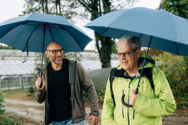 Zwei Männer mit Brille spazieren lachend unter blauen Regenschirmen auf einem Weg neben Gewächshäusern und Bäumen. Einer trägt eine grüne Outdoorjacke und Rucksack, der andere eine braune Jacke.