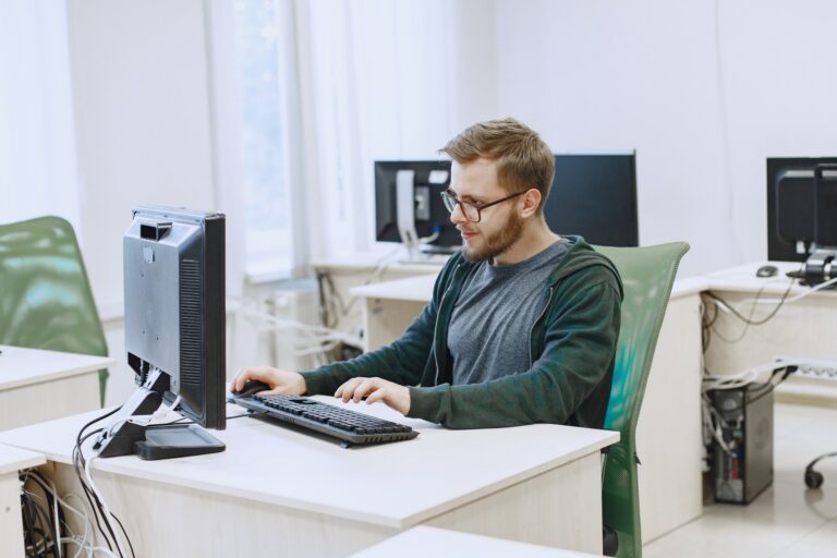 Junger Mann mit Brille und Bart sitzt in einem hellen Büroraum an einem Schreibtisch und arbeitet konzentriert am Computer. Im Hintergrund stehen weitere leere Arbeitsplätze mit Bildschirmen.